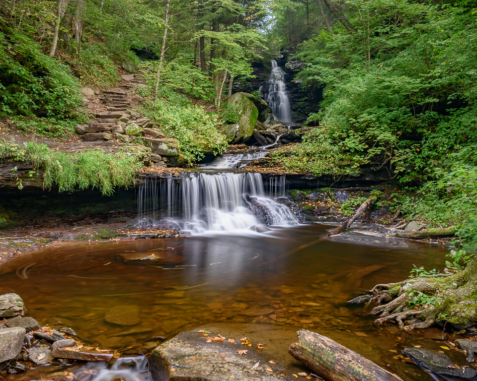 A trail runs alongside a waterfall in the woods.