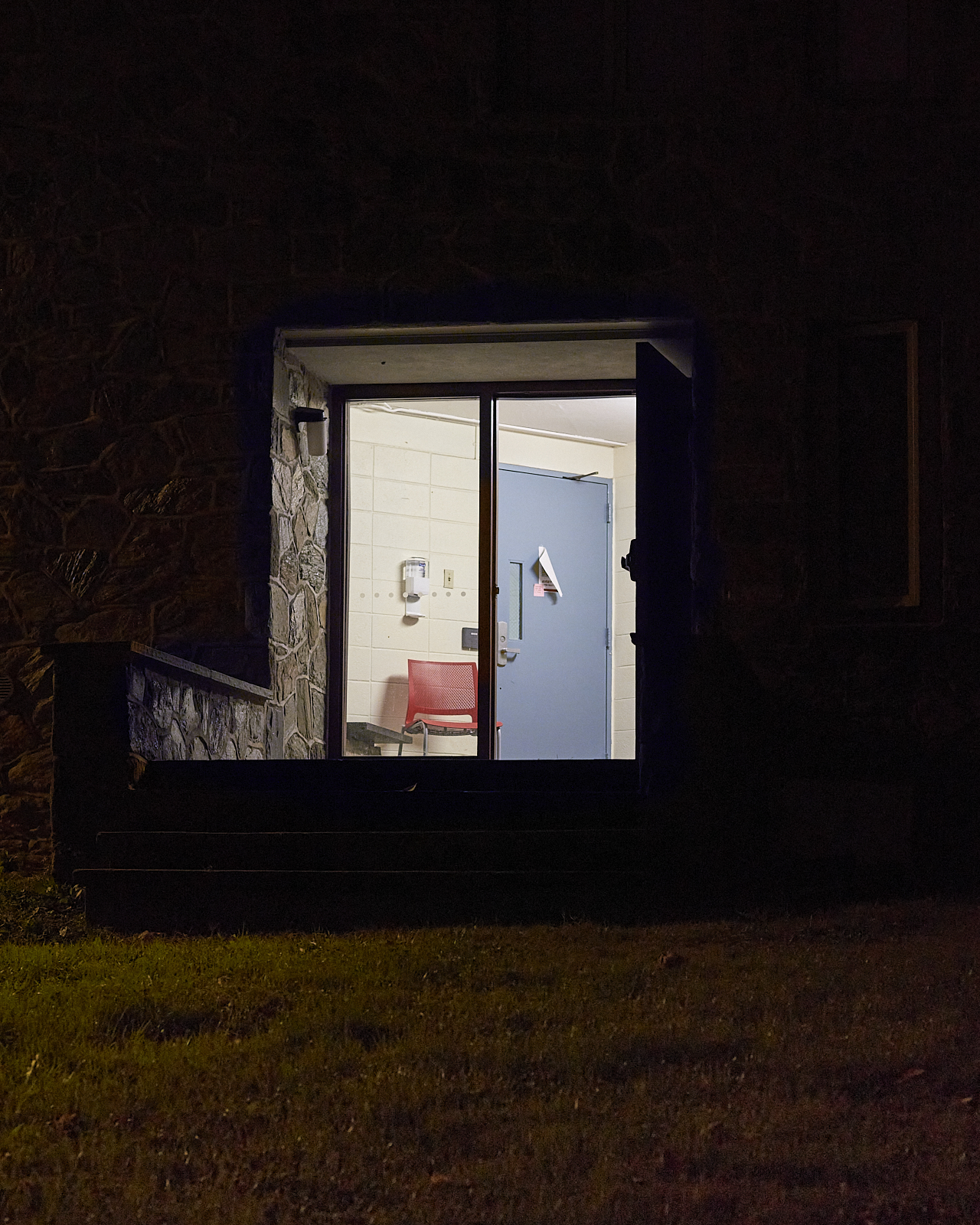 Urban #221007.2 A color photograph of an entryway at night. Inside a red plastic chair sits next to a blue metal door. A hand sanitizer hangs on the cream-colored cinderblock wall.