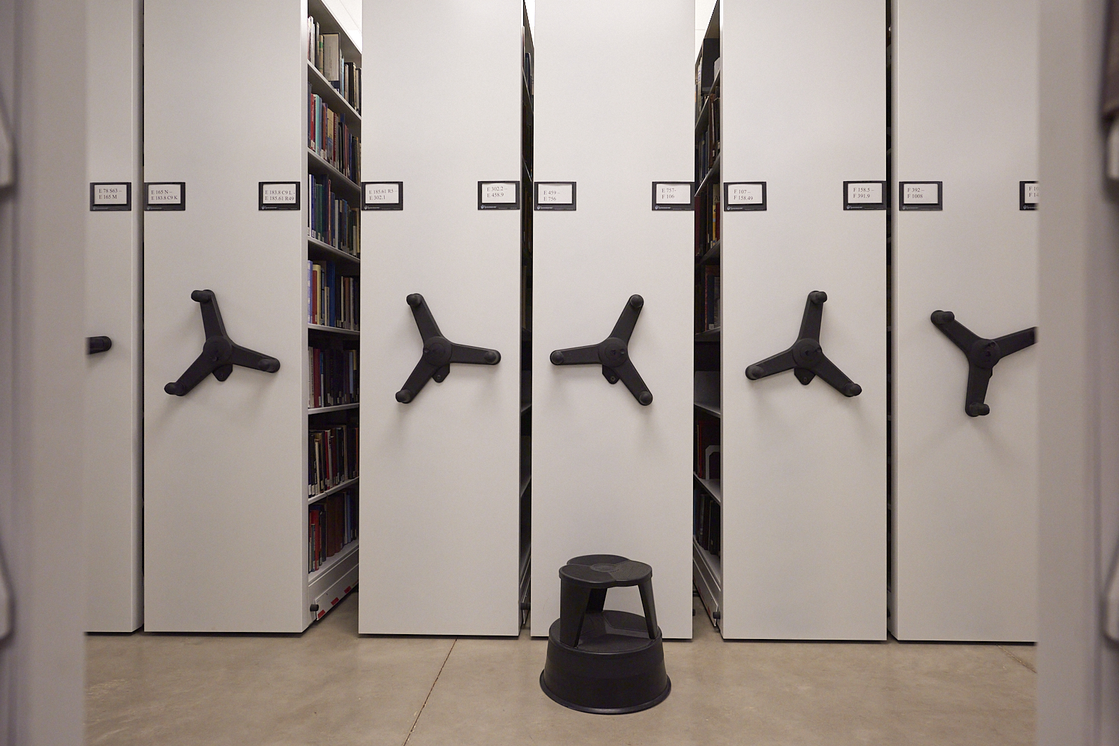 Interior #220830. A color photograph of the ends of compact shelving, with a stool on the floor in front of them.