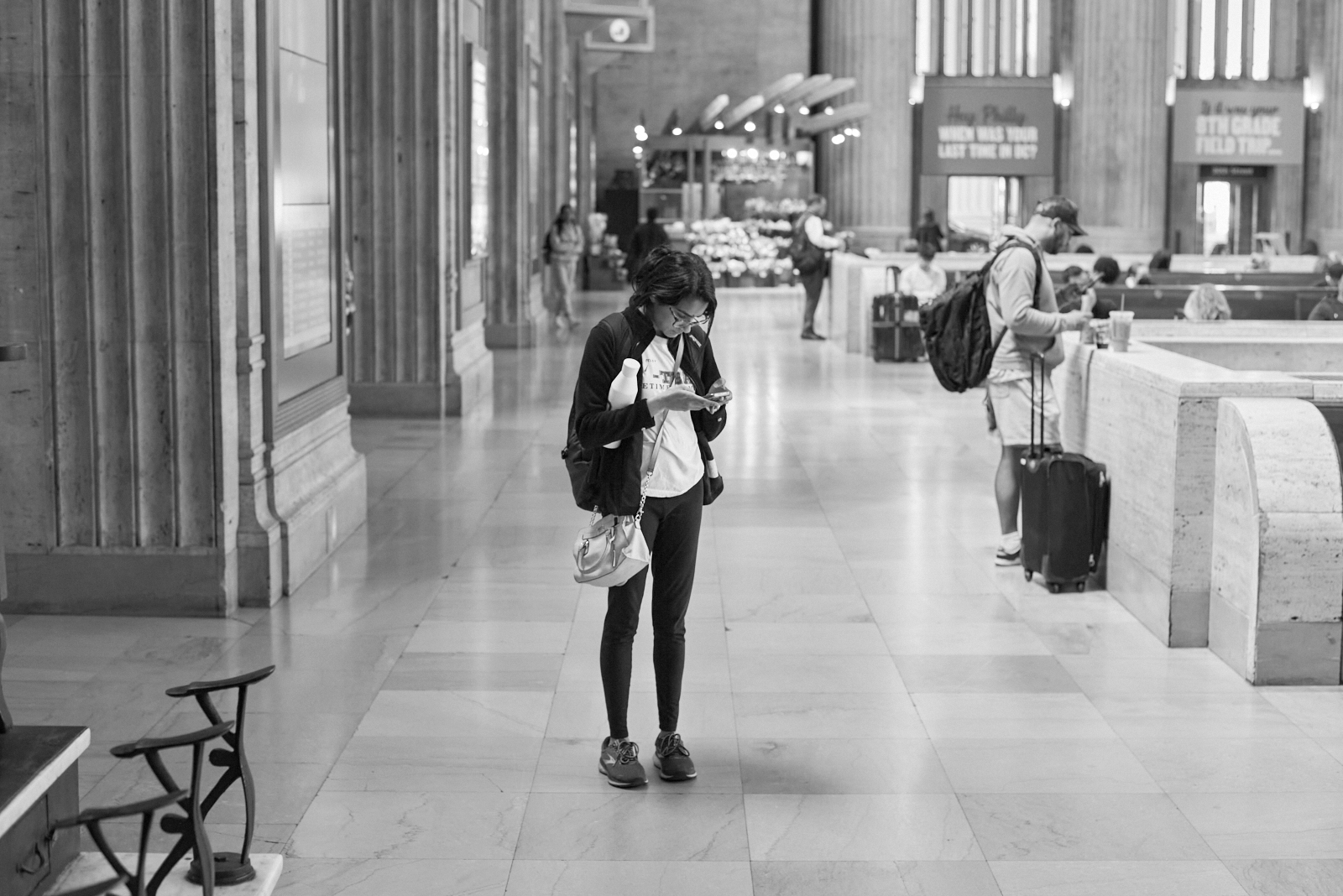 Urban #230510.2. A black and white photo of a woman standing in 30th Street Station looking at her phone.