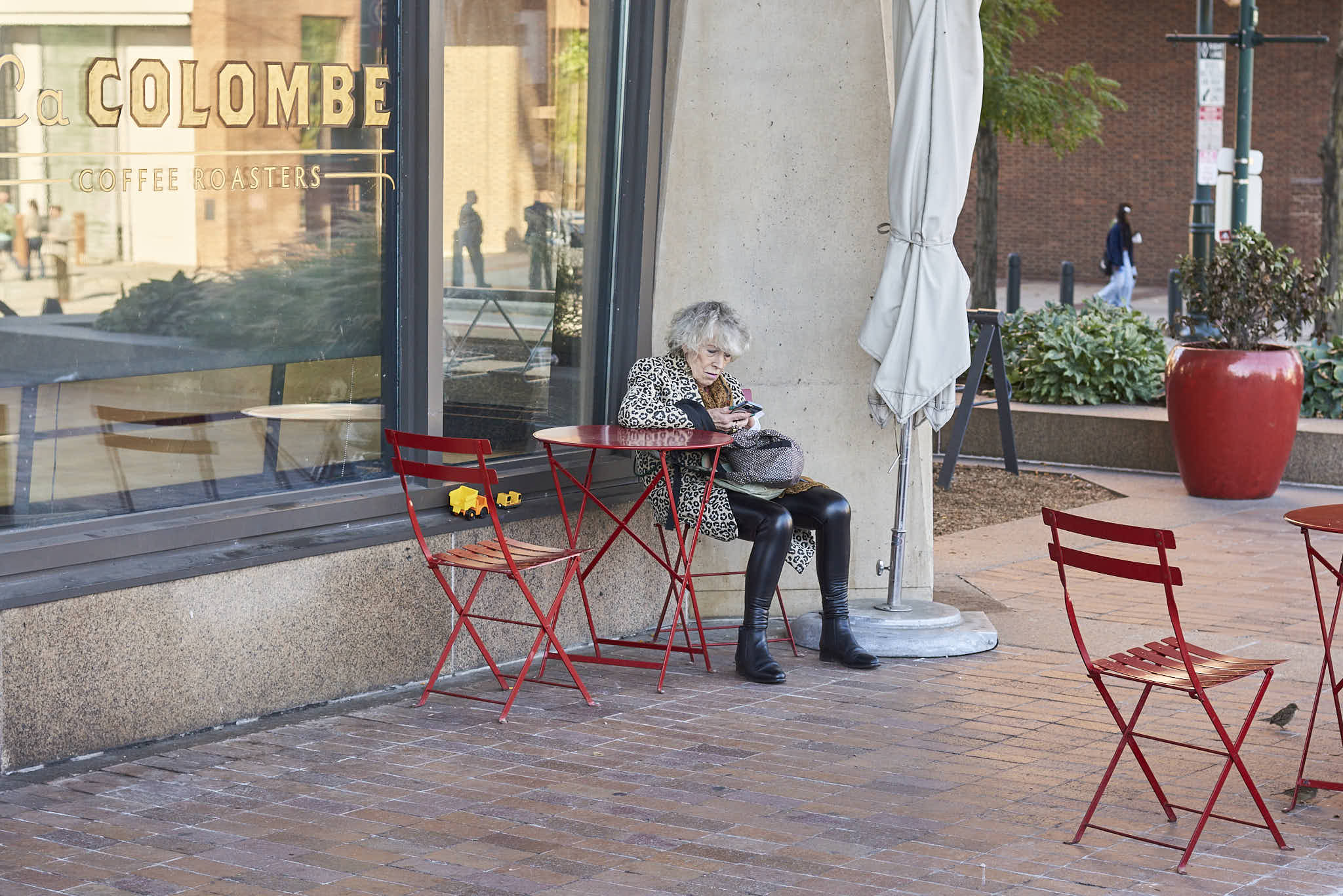 Urban #231013.5. A color photo of a woman sitting at a red metal table outside a cafe.