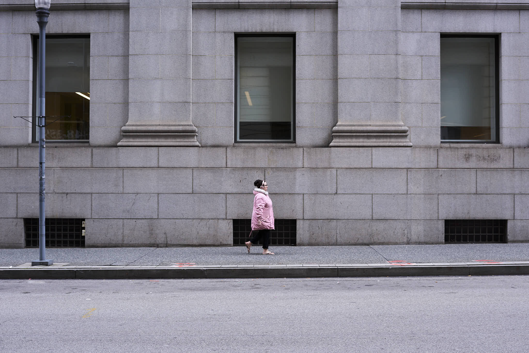 Urban #231015. Color photograph of a woman wearing a pink coat and headphones and flip flops walking up a street in front of a grey building.