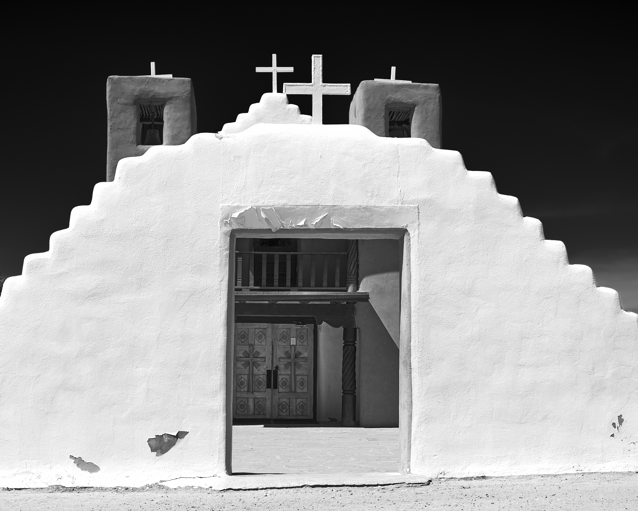 Urban #230820. A black and white photo of a bright white wall in front of a small adobe church. The sky is dark, though it is midday.