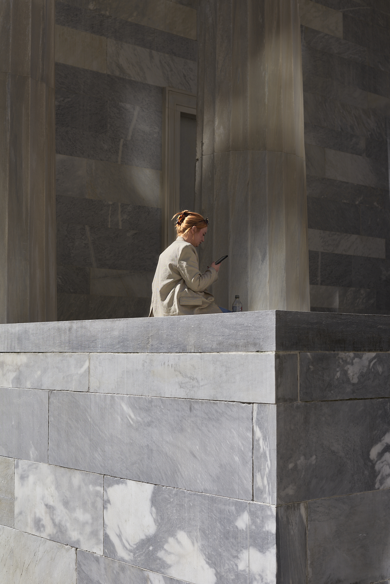 Urban #231013.4. Color photograph of a woman sitting on a stone ledge in front of a building. She is looking at her phone.