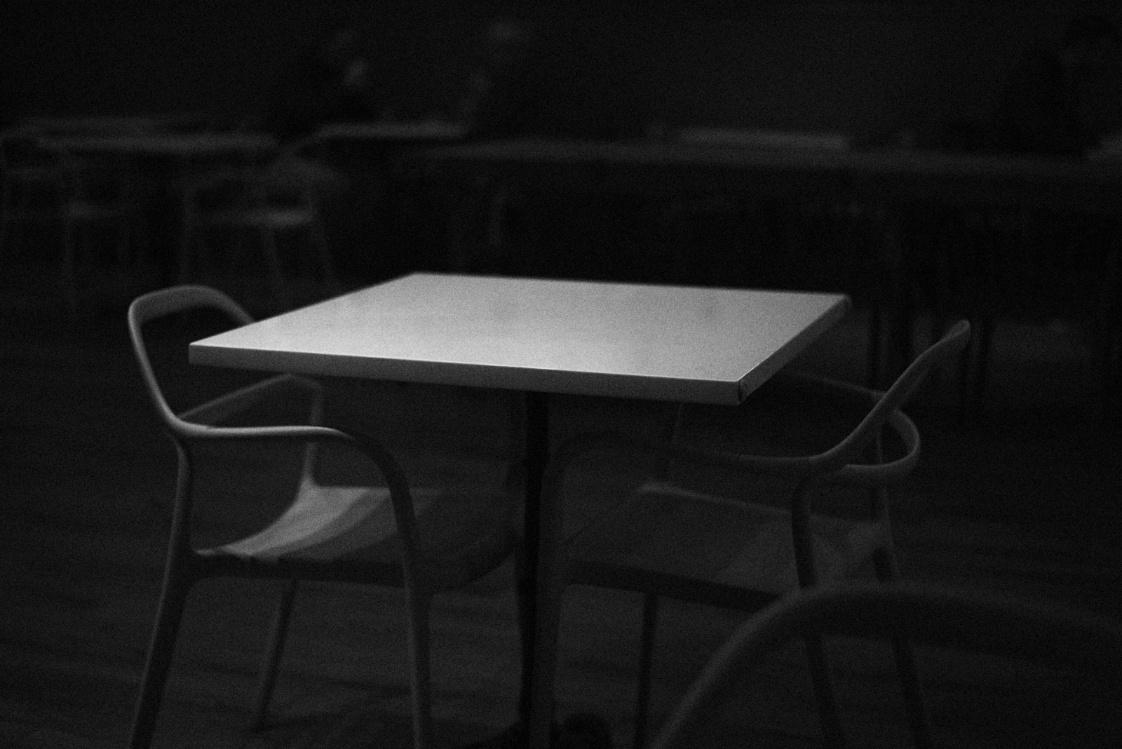 Urban #231223. A black and white photo of a table with two chairs in a darkened space.