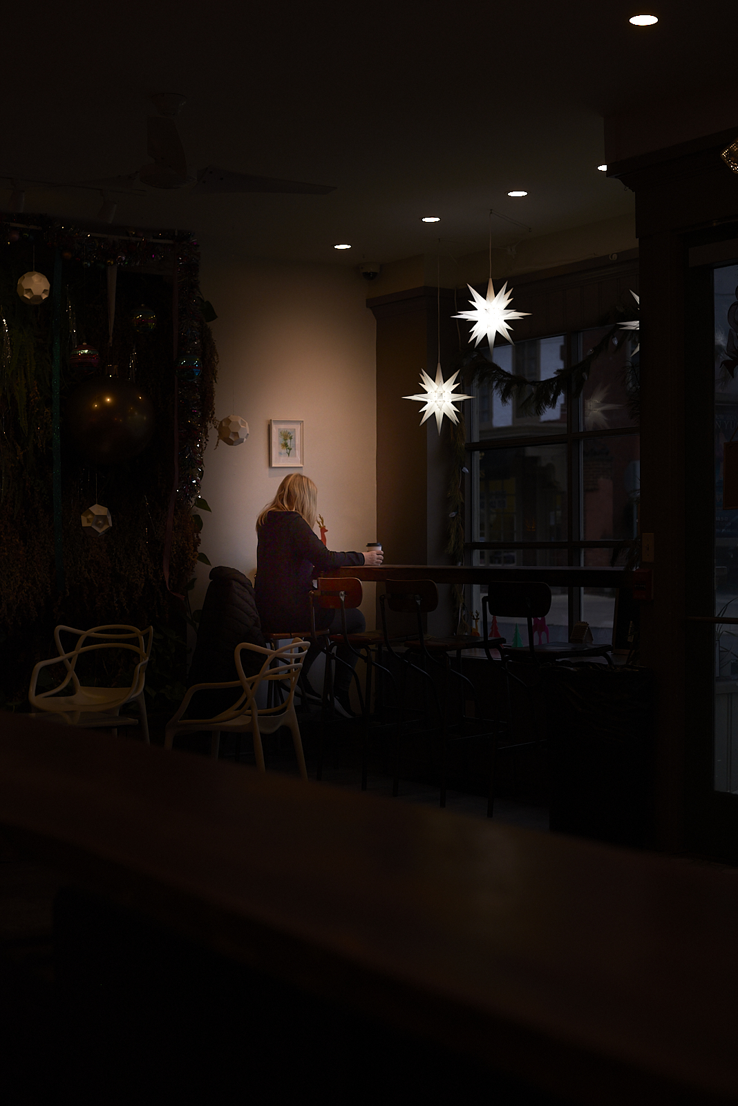 Urban #231223.1. A color photograph of a woman sitting alone by a window, two lighted stars hang above her head.