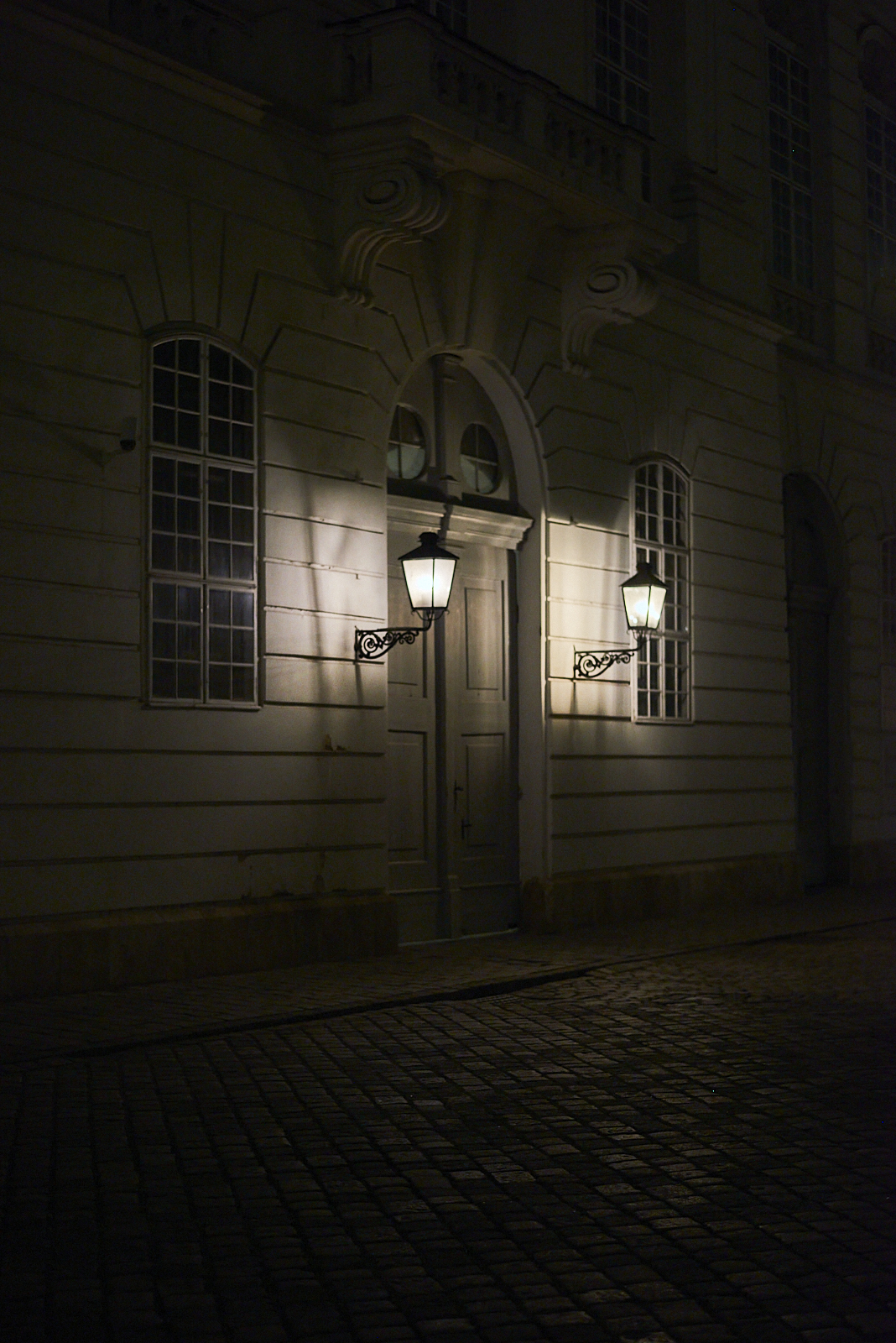 Urban #240309.2. A dark photograph of a doorway, two lights on either side and some cobblestones in front.