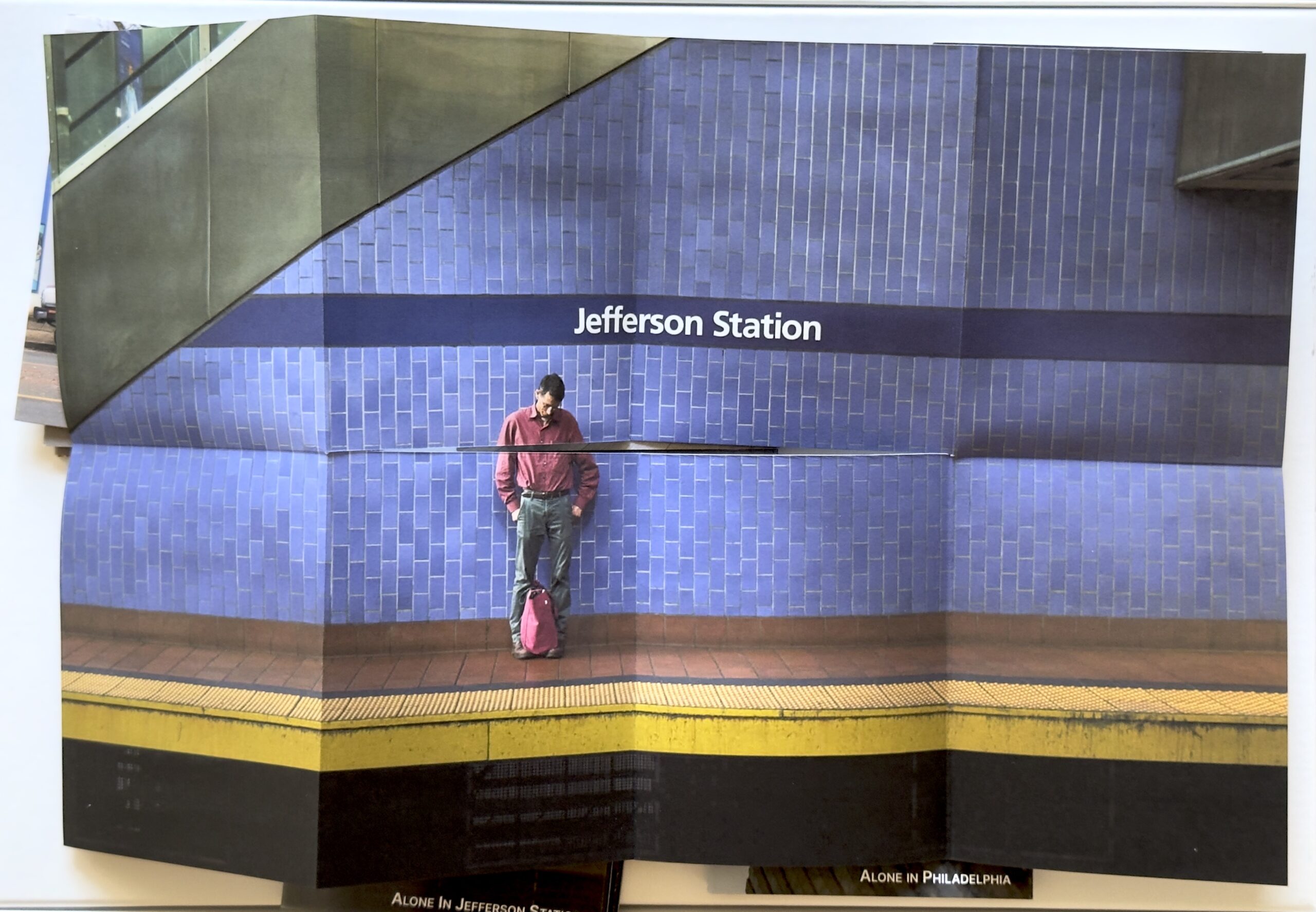 Color photograph of the central image in the “Alone in Jefferson” zine. A man stands alone against a blue tiled wall. He looks towards his feet.