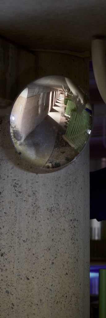 Urban #250417.2 is a color photograph of a pillar and a circular mirror. The mirror is at the top of the frame and reflects a series of lights down the hallway.